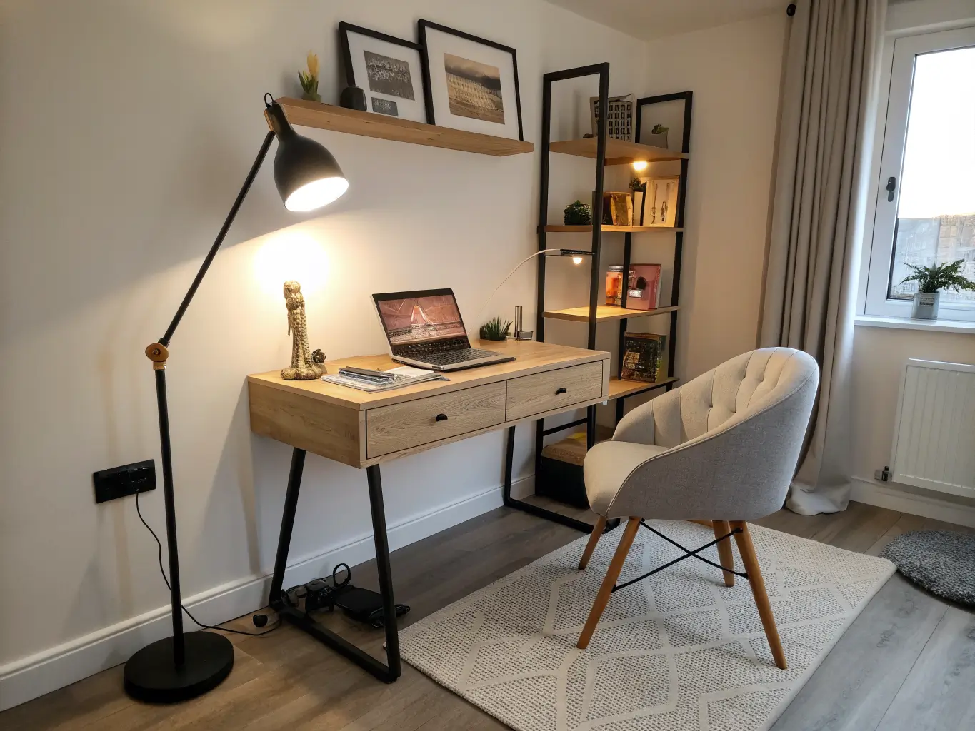 Wide-angle view from a doorway of a bedroom study corner at dusk with a light oak desk, laptop, layered warm lighting, and a chair with clear walkway space.