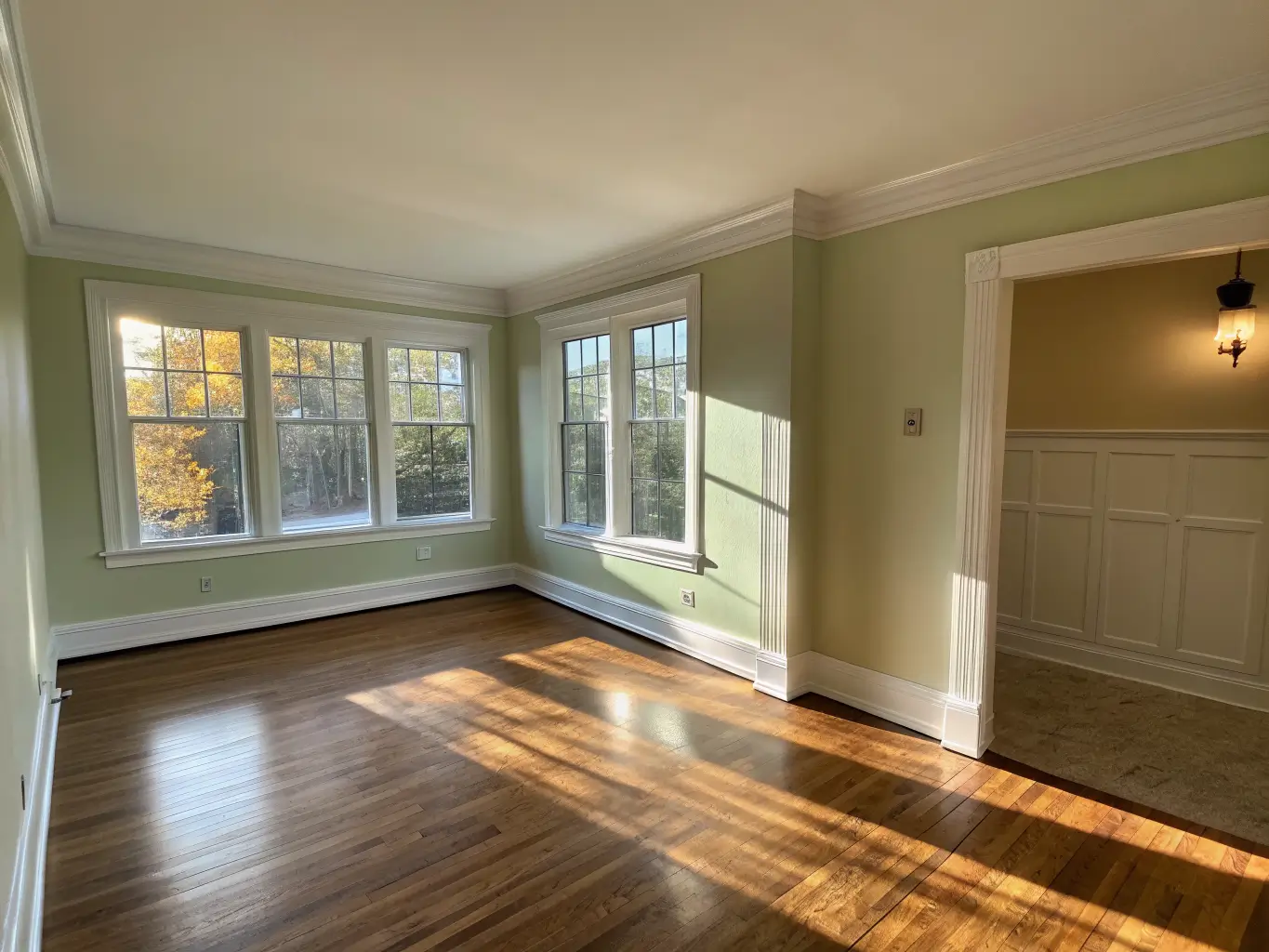 Wide-angle photo of an empty living room corner in late-afternoon sunlight, with fresh sage-green paint on the back wall and oak floors showing a dust-free rectangle.