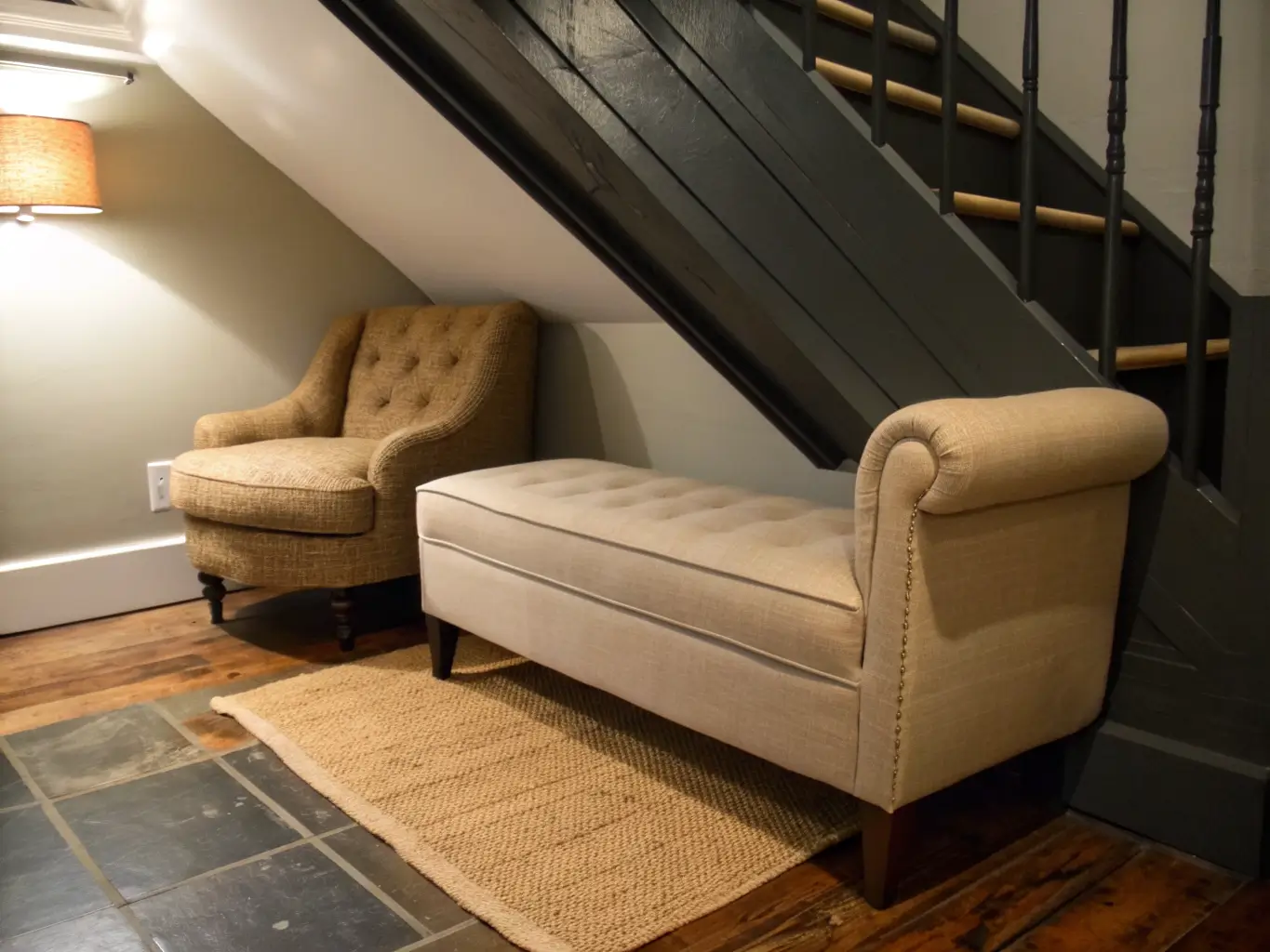 Photorealistic under-stairs reading nook with oatmeal linen bench on plywood platform, low rolled-arm chair, jute rug, and warm brass picture light under a sloped charcoal ceiling at dusk.